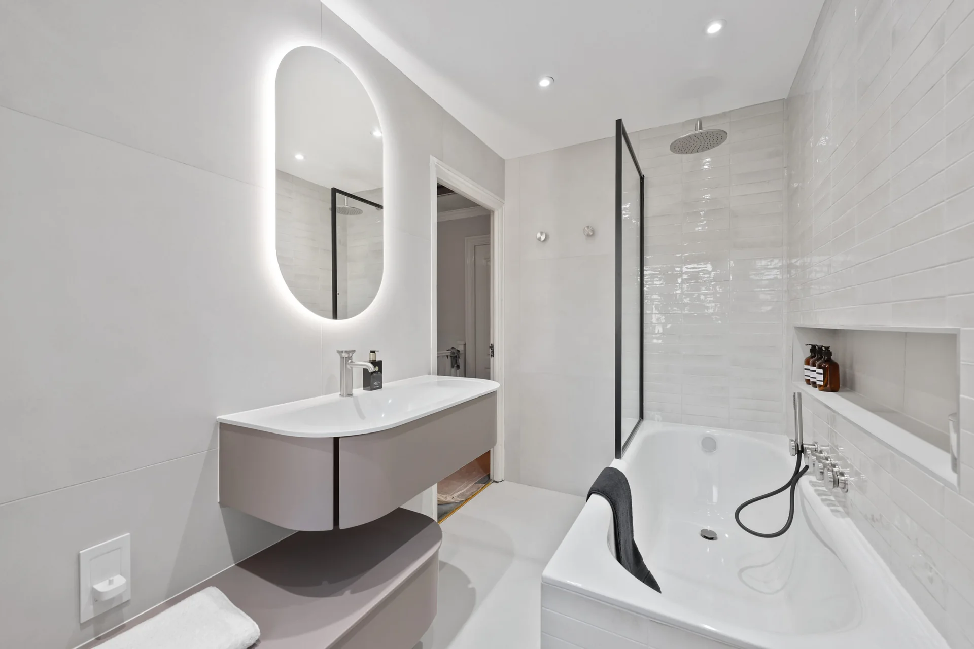 Completed bath-and-shower room with a curved floating vanity, asymmetric mirror, recessed niche and black-edged screen.