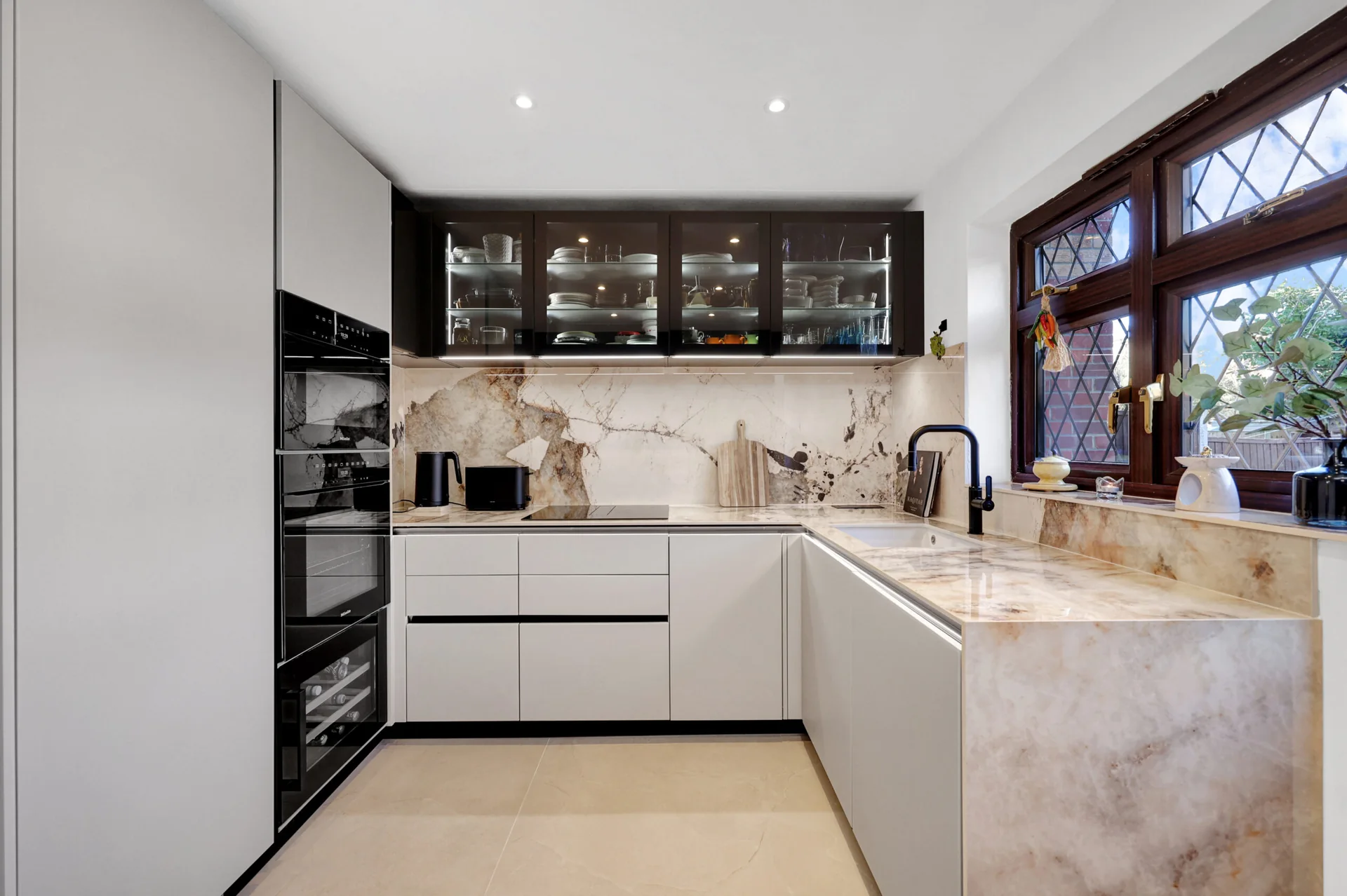 Completed handleless kitchen with white furniture, dark glazed wall cabinets, strongly veined stone surfaces and retained timber windows.